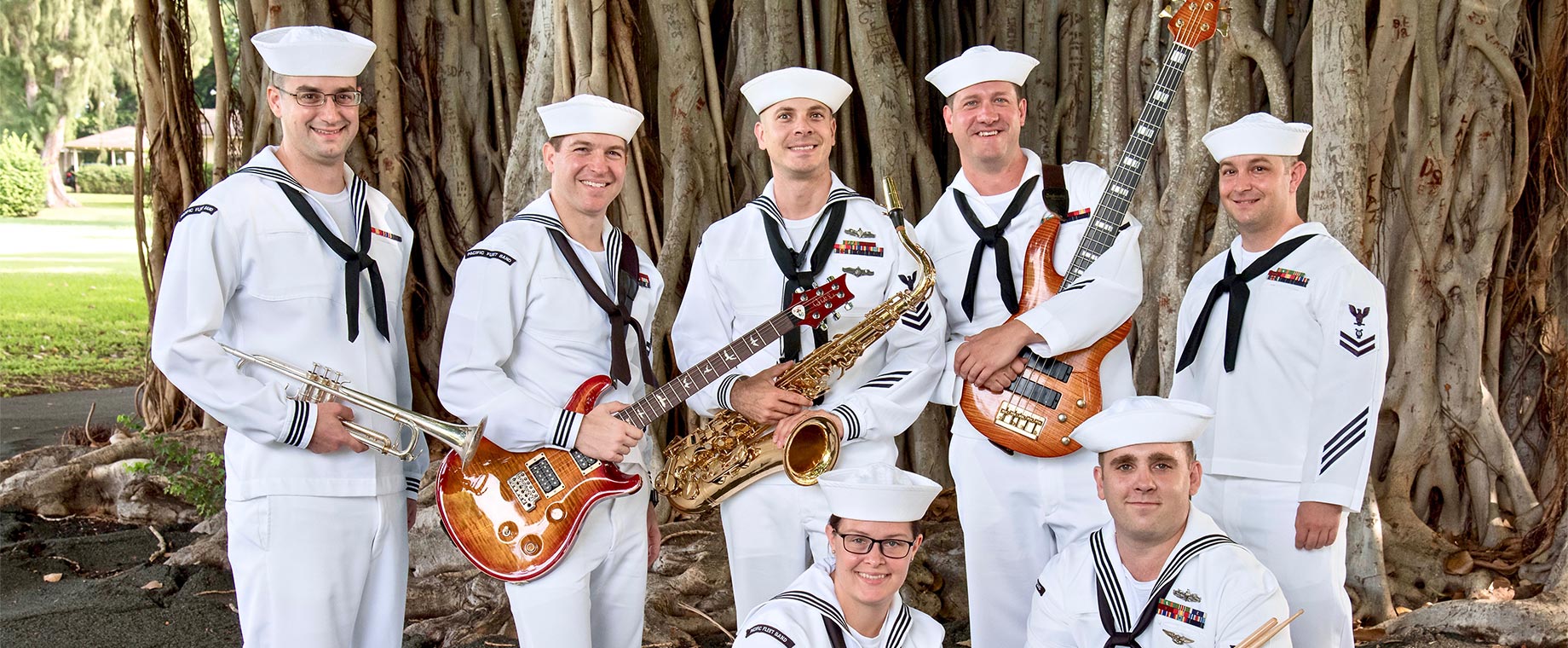 Seven Sailors smile for the camera. Five are holding musical instruments: a trumpet, electric guitar, saxophone, bass guitar and drum sticks.