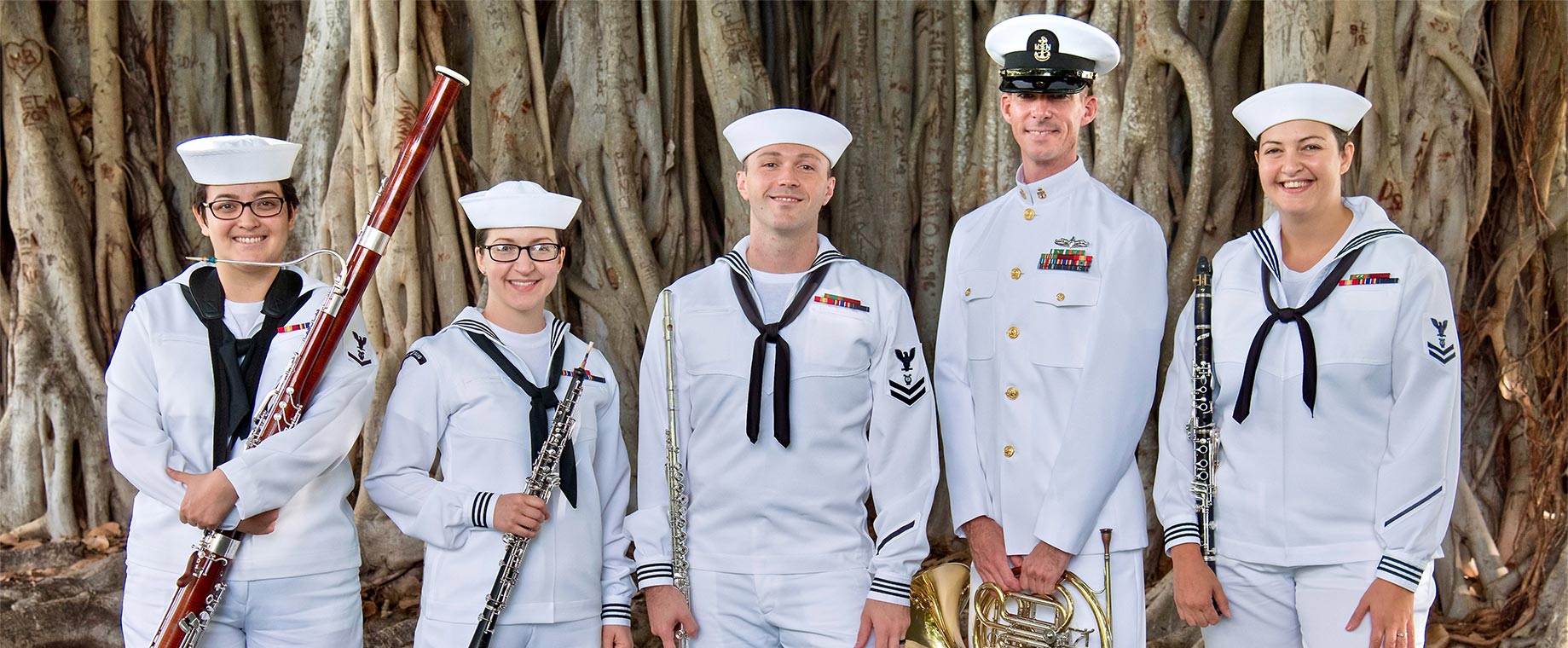 Five Sailors smile at the camera while holding their instruments: a bassoon, oboe, flute, french horn and clarinet.